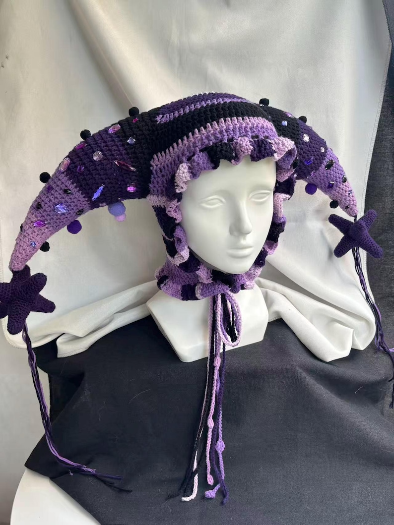 Pode incluir: Um gorro de bobo da corte em croch&ecirc; nos tons roxo escuro, roxo claro e preto. O gorro apresenta dois pain&eacute;is laterais longos e pontiagudos adornados com pequenos pompons pretos e v&aacute;rias contas facetadas roxas e transparentes. Uma estrela pendura.