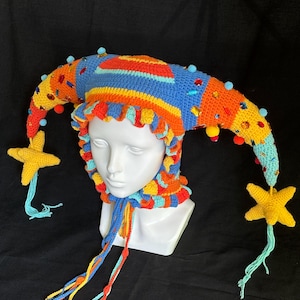 Pode incluir: Um gorro de bobo da corte em croch&ecirc; multicolorido com duas pontas longas e curvas que terminam em borlas amarelas em forma de estrela. O gorro &eacute; decorado com pequenos pompons azuis, gemas vermelhas e amarelas e um babado com la&ccedil;os longos.