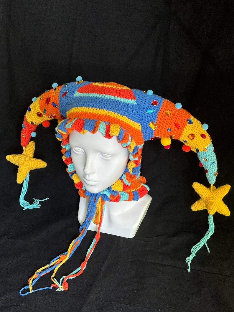 Pode incluir: Um gorro de bobo da corte em croch&ecirc; multicolorido com duas pontas longas e curvas que terminam em borlas amarelas em forma de estrela. O gorro &eacute; decorado com pequenos pompons azuis, gemas vermelhas e amarelas e um babado com la&ccedil;os longos.