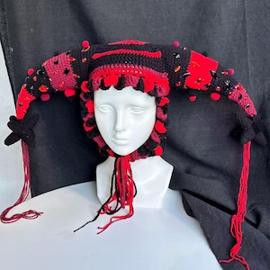 Pode incluir: Um gorro de bobo da corte em croch&ecirc; com listras alternadas em preto e vermelho, dois chifres laterais longos e pontiagudos e pompons vermelhos decorativos. O gorro &eacute; adornado com cristais facetados vermelhos e pretos e termina com detalhes em forma de estrela preta e longas borlas de fio vermelho.