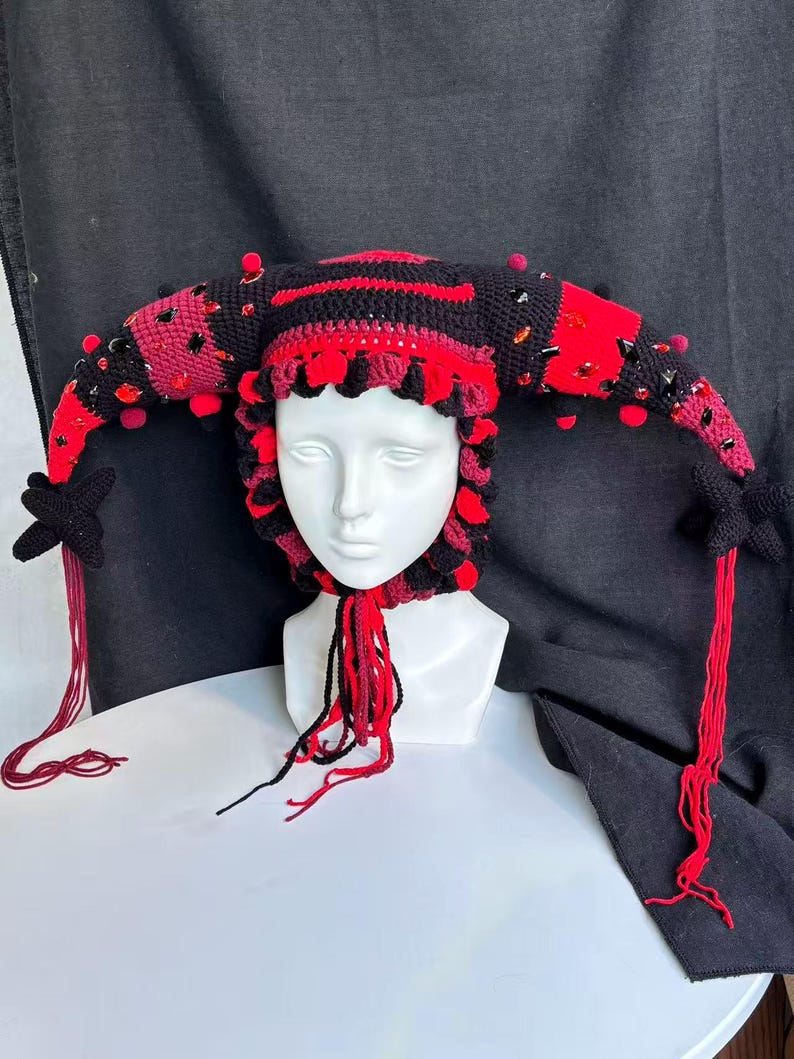 Pode incluir: Um gorro de bobo da corte em croch&ecirc; com listras alternadas em preto e vermelho, dois chifres laterais longos e pontiagudos e pompons vermelhos decorativos. O gorro &eacute; adornado com cristais facetados vermelhos e pretos e termina com detalhes em forma de estrela preta e longas borlas de fio vermelho.
