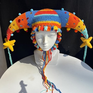 Pode incluir: Um chap&eacute;u de bobo da corte em croch&ecirc; multicolorido com dois chifres longos e curvos adornados com pequenos pompons azuis, strass coloridos e pingentes em forma de estrela amarela nas pontas. O chap&eacute;u tem uma borda franzida e la&ccedil;os tran&ccedil;ados.