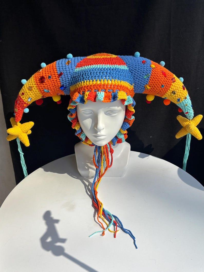 Pode incluir: Um chap&eacute;u de bobo da corte em croch&ecirc; multicolorido com dois chifres longos e curvos adornados com pequenos pompons azuis, strass coloridos e pingentes em forma de estrela amarela nas pontas. O chap&eacute;u tem uma borda franzida e la&ccedil;os tran&ccedil;ados.
