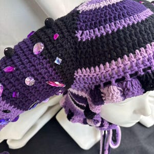 Pode incluir: Um gorro de croch&ecirc; com uma se&ccedil;&atilde;o de cauda longa e pontiaguda, feito em listras alternadas de fio preto, roxo escuro e roxo claro. O gorro &eacute; decorado com pequenos pompons pretos e v&aacute;rios strass acr&iacute;licos facetados roxos e transparentes.