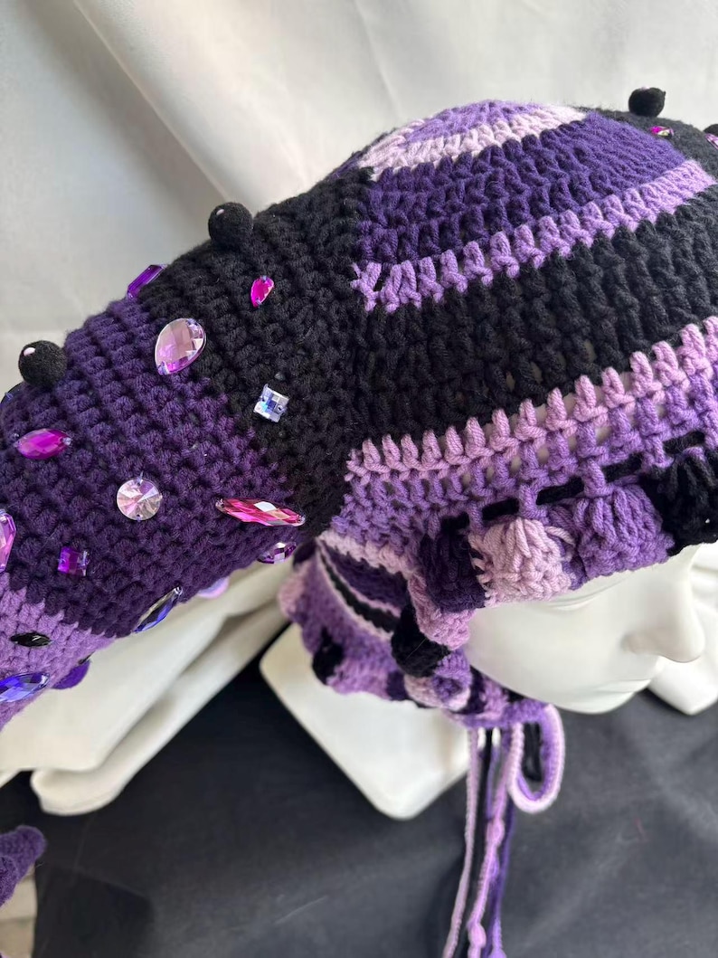 Pode incluir: Um gorro de croch&ecirc; com uma se&ccedil;&atilde;o de cauda longa e pontiaguda, feito em listras alternadas de fio preto, roxo escuro e roxo claro. O gorro &eacute; decorado com pequenos pompons pretos e v&aacute;rios strass acr&iacute;licos facetados roxos e transparentes.