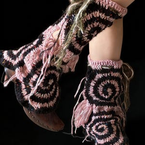 May include: Crocheted leg warmers and ankle cuffs in black and pink spiral patterns. The leg warmers have fringe and are worn over wooden clogs. The cuffs have a pink trim and are worn around the ankles.
