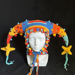 Pode incluir: Um chap&eacute;u de bobo da corte em croch&ecirc; multicolorido com dois chifres longos e curvos adornados com pequenos pompons azul-claros, strass coloridos e pingentes em forma de estrela amarela nas pontas. O chap&eacute;u tem uma borda franzida e la&ccedil;os tran&ccedil;ados.
