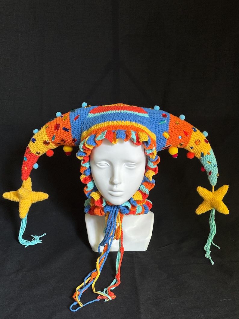 Pode incluir: Um chap&eacute;u de bobo da corte em croch&ecirc; multicolorido com dois chifres longos e curvos adornados com pequenos pompons azul-claros, strass coloridos e pingentes em forma de estrela amarela nas pontas. O chap&eacute;u tem uma borda franzida e la&ccedil;os tran&ccedil;ados.