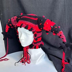 Pode incluir: Um gorro de bobo da corte em croch&ecirc; com listras horizontais alternadas em preto e vermelho, adornado com cristais facetados vermelhos e pretos espalhados. O gorro tem duas extens&otilde;es laterais longas e pontiagudas terminadas em borlas de estrela.