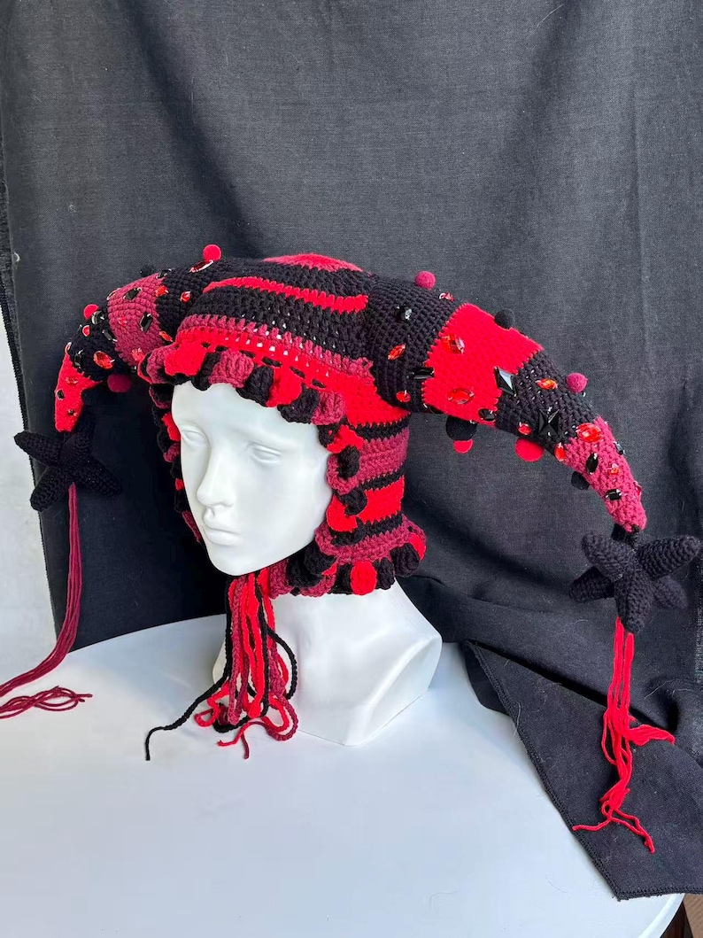 Pode incluir: Um gorro de bobo da corte em croch&ecirc; com listras horizontais alternadas em preto e vermelho, adornado com cristais facetados vermelhos e pretos espalhados. O gorro tem duas extens&otilde;es laterais longas e pontiagudas terminadas em borlas de estrela.