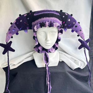 Pode incluir: Um gorro de bobo da corte em croch&ecirc; nos tons preto e roxo, com dois chifres longos e curvos adornados com pequenos pompons escuros e cristais roxos e transparentes. O gorro tem uma borda com babados e dois pingentes em forma de estrela.