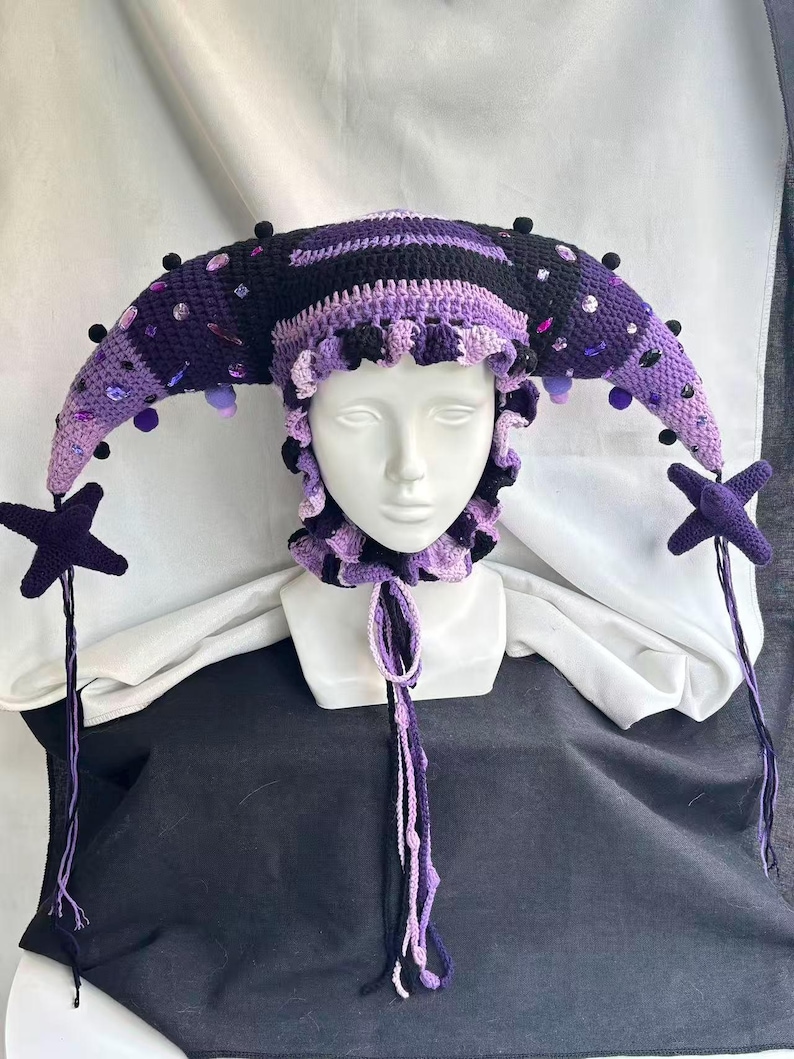 Pode incluir: Um gorro de bobo da corte em croch&ecirc; nos tons preto e roxo, com dois chifres longos e curvos adornados com pequenos pompons escuros e cristais roxos e transparentes. O gorro tem uma borda com babados e dois pingentes em forma de estrela.