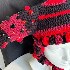 Pode incluir: Um gorro de croch&ecirc; com um padr&atilde;o de listras pretas e vermelhas e duas estruturas salientes em forma de chifre. O gorro &eacute; adornado com numerosas pedras preciosas facetadas vermelhas e pretas e pequenos pompons, com la&ccedil;os tran&ccedil;ados pendentes.