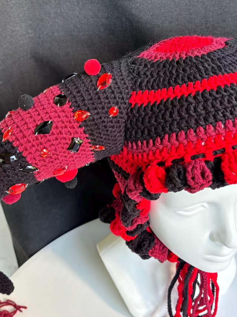 Pode incluir: Um gorro de croch&ecirc; com um padr&atilde;o de listras pretas e vermelhas e duas estruturas salientes em forma de chifre. O gorro &eacute; adornado com numerosas pedras preciosas facetadas vermelhas e pretas e pequenos pompons, com la&ccedil;os tran&ccedil;ados pendentes.