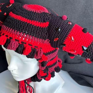 Pode incluir: Um gorro de croch&ecirc; preto e vermelho com duas proje&ccedil;&otilde;es proeminentes em forma de chifre, adornado com pequenos pompons vermelhos e cristais facetados vermelhos e pretos. O gorro tem um padr&atilde;o listrado texturizado e borlas de fio pendentes.