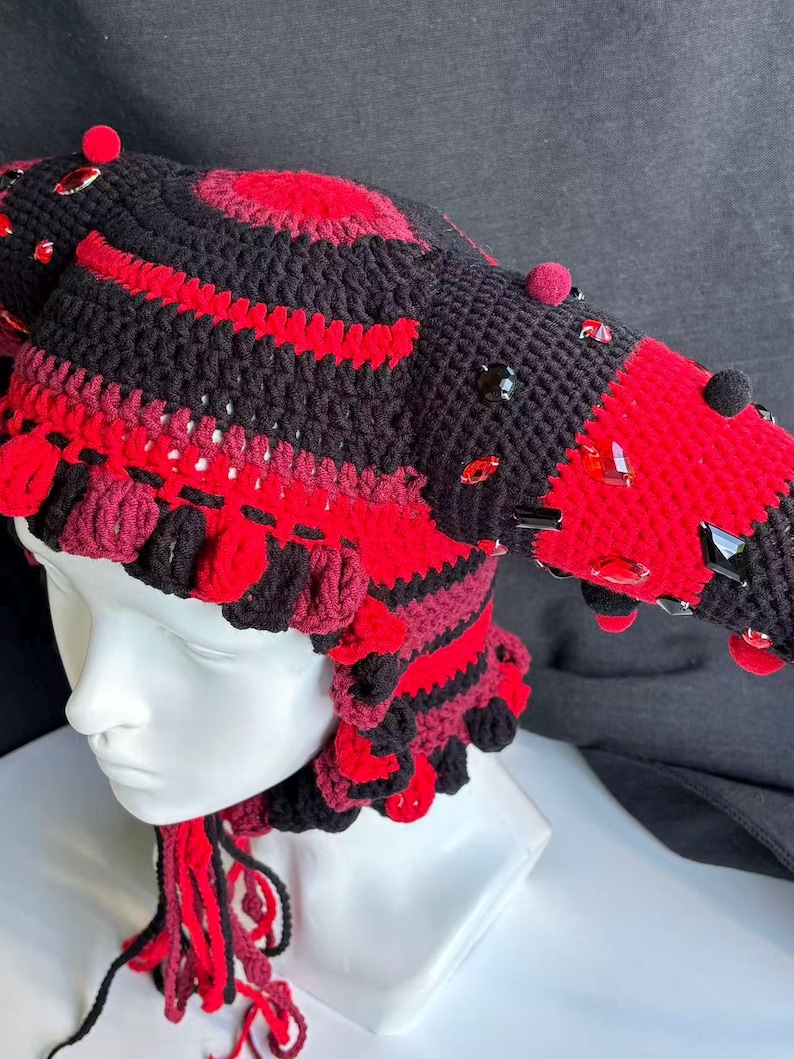 Pode incluir: Um gorro de croch&ecirc; preto e vermelho com duas proje&ccedil;&otilde;es proeminentes em forma de chifre, adornado com pequenos pompons vermelhos e cristais facetados vermelhos e pretos. O gorro tem um padr&atilde;o listrado texturizado e borlas de fio pendentes.