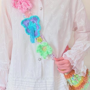 May include: A white eyelet shirt is adorned with a colorful, handmade crochet floral accessory. The accessory features pink, blue, and green flowers, and a small, decorative purse with a beaded handle.