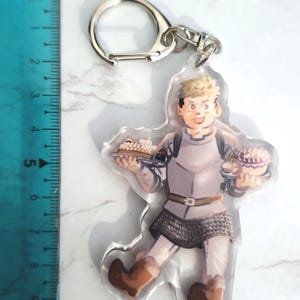 May include: A clear acrylic keychain with a cartoon illustration of a person in medieval clothing, carrying plates of food. The keychain has a silver metal ring and clasp. The ruler next to the keychain measures up to 25 cm.