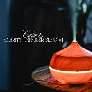 Puede incluir: Un difusor de aceites esenciales naranja y blanco con el texto "Calm & Clarity Diffuser Blend #1". El difusor emite un chorro de vapor. El difusor está sobre una mesa de madera.