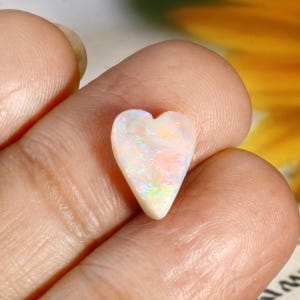 Puede incluir: Una piedra preciosa de ópalo en forma de corazón, con un color base blanco nacarado y destellos iridiscentes. La piedra se sostiene en una mano, mostrando su superficie pulida y su juego de colores único. El ópalo es una piedra natural.