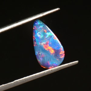 May include: A teardrop-shaped opal gemstone, held by tweezers, displays a vibrant play of color. The stone's surface features a mix of blue, red, and orange hues, creating a mesmerizing effect against a dark background. The opal is a natural gemstone.