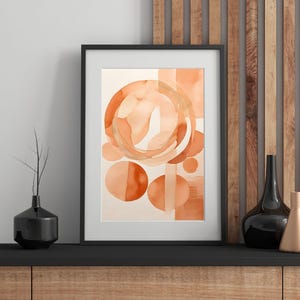 Op de afbeelding: Ingelijste abstracte aquarel kunstdruk met oranje en perzikkleurige cirkels en vormen. Het kunstwerk wordt gepresenteerd in een zwart frame. Decoratieve kunst voor woondecoratie.