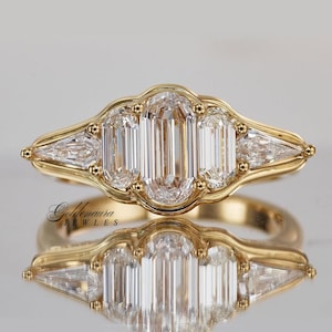 Puede incluir: Un anillo de oro con tres diamantes de talla esmeralda, flanqueados por dos diamantes triangulares. El anillo tiene un engaste ornamentado con pequeños detalles dorados. El anillo se muestra sobre una superficie reflectante.