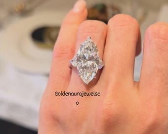 Könnte beinhalten: Ein silberfarbener Ring mit einem großen, marquise-geschliffenen Mittelstein, der von zwei kleineren, birnenförmigen Seitensteinen flankiert wird. Der Text Goldenaurajewelsc ist schwarz auf der Haut neben dem Ring aufgedruckt. Die Fassung ist metallisch.