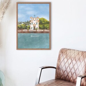 May include: A watercolour painting of a white church with a cross, set against a blue sky and green foliage. The church is reflected in the turquoise water below. The artwork is framed in a wooden frame.