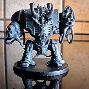 May include: A gray plastic miniature figure of a heavily armored, monstrous humanoid. It features a skull-like face, horns, and multiple weapon attachments, including a double-barreled cannon. The figure is mounted on a black circular base.