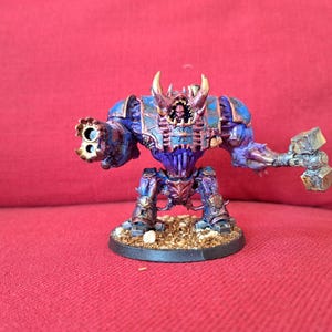 May include: A detailed miniature figure of a robotic warrior, predominantly blue and purple with metallic accents. It features a multi-barreled weapon on one arm and a hammer on the other. The figure stands on a textured base.