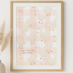 May include: A framed print with a repeating pattern of white geese set against a pink and white gingham backdrop. The print includes the text "Linger where the light is kind". The frame is made of light-coloured wood.