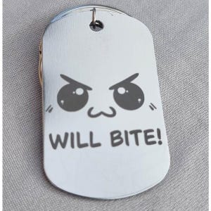 Könnte beinhalten: Silberfarbene Hundemarke mit einem Cartoon-Gesicht und dem Text "WILL BITE!" in Schwarz. Die Marke hat oben einen kleinen Ring zur Befestigung.