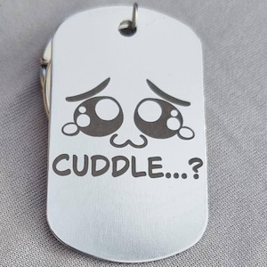 Könnte beinhalten: Eine silberfarbene Hundemarke mit abgerundeter rechteckiger Form. Die Marke zeigt ein eingraviertes Design mit großen Augen im Cartoon-Stil und dem Wort "CUDDLE...?" in einer verspielten Schriftart. Oben ist ein kleiner Metallring angebracht.