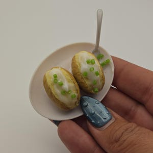 Könnte beinhalten: Ein ovaler Miniaturteller in Weiß mit zwei kleinen braunen Ofenkartoffeln, die mit weißer Creme und grünen Schnittlauchröllchen belegt sind, dazu ein winziger weißer Löffel. Die Speisen sind texturiert für ein realistisches Aussehen.