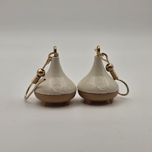 May include: A pair of drop earrings featuring miniature white, bell-shaped charms with a small power button icon embossed on the front. The charms have a tan base with three tiny feet and are attached to gold-toned metal ear wires with a coiled detail.