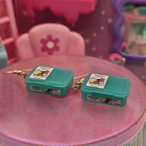May include: A pair of dangle earrings featuring miniature teal plastic lunch boxes. Each rectangular container has a hinged lid with a label displaying a sandwich and the text "sistema lunch". The earrings are attached to gold-tone metal hooks.