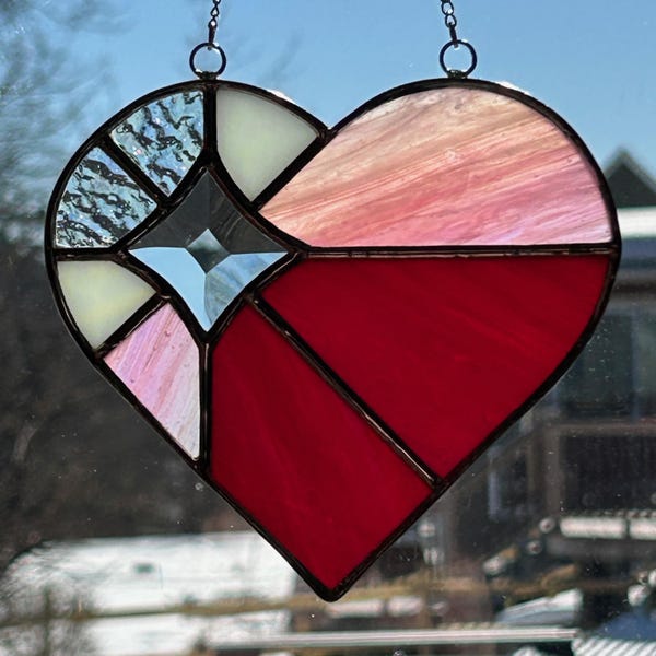 May include: Stained glass heart suncatcher with a black metal frame. The heart is divided into sections of red, pink, white, and blue glass. The suncatcher hangs from a chain. The heart shape is a symbol of love and affection.