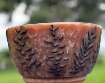 May include: A bowl with a marbled, translucent amber and brown resin finish featuring dark, etched pine tree silhouettes around the exterior. The bowl is mounted on a light-colored, turned wooden pedestal base with a smooth, cylindrical shape.