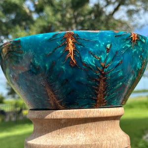 May include: A bowl crafted from a combination of natural wood and translucent teal resin. The design features intricate, branch-like wood inclusions embedded within the vibrant, swirling blue-green resin, creating a unique, organic pattern.