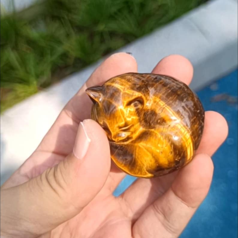 May include: A carved tiger's eye gemstone shaped like a sleeping cat. The polished stone features bands of golden brown and dark brown, with the cat's features delicately carved. The cat is curled up in a ball, with its head resting on its paws.