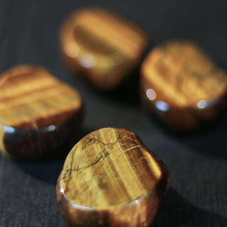 May include: Four polished tiger's eye stones. The stones are a mix of golden brown and dark brown, with a glossy finish. The stones have a smooth, rounded shape and are arranged on a dark surface.