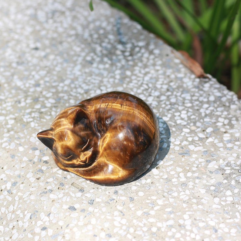 May include: A polished, brown tiger's eye gemstone carved into the shape of a curled-up sleeping cat. The stone has a natural, striped pattern with shades of gold and brown. The cat figurine is resting on a speckled, gray surface.