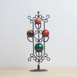 Op de afbeelding: Een zwarte metalen displaystandaard met vijf kleurrijke edelsteenbollen. De bollen zijn rood, groen en bruin, en de standaard heeft een decoratief, krullend ontwerp. De standaard staat op een houten oppervlak.