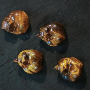 May include: Four polished tiger's eye stone carvings, each shaped like a small bird. The stones display a rich, golden-brown color with natural streaks and patterns. The carvings are set against a dark, textured background.