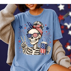 May include: Light blue t-shirt featuring a skeleton wearing sunglasses and a headband with an American flag design. The skeleton is holding a beer mug with an American flag design and making a peace sign. Fireworks and stars are in the background.