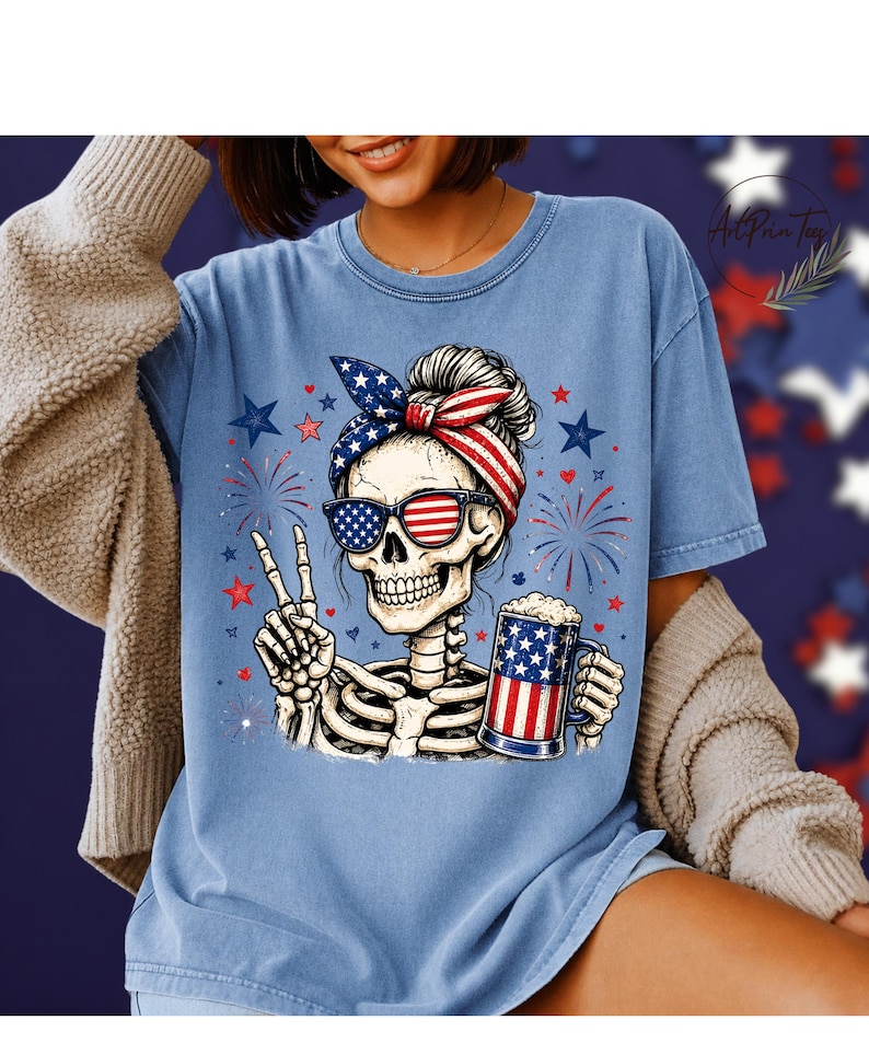 May include: Light blue t-shirt featuring a skeleton wearing sunglasses and a headband with an American flag design. The skeleton is holding a beer mug with an American flag design and making a peace sign. Fireworks and stars are in the background.