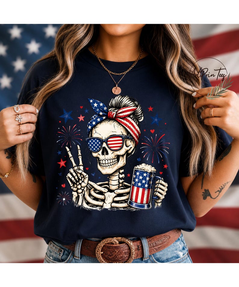 May include: Navy blue t-shirt featuring a skeleton wearing sunglasses and a headband with an American flag design. The skeleton is holding a beer mug with an American flag design and making a peace sign. Fireworks and stars are in the background.