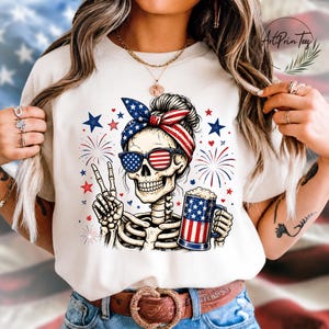 May include: A cream-colored t-shirt featuring a skeleton with sunglasses and a patriotic headband. The skeleton is holding a beer mug and making a peace sign. Fireworks and stars are in the background.