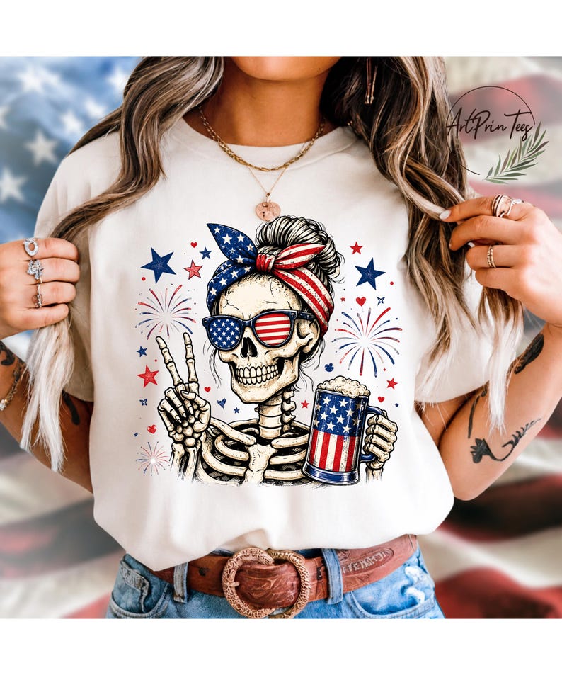 May include: A cream-colored t-shirt featuring a skeleton with sunglasses and a patriotic headband. The skeleton is holding a beer mug and making a peace sign. Fireworks and stars are in the background.